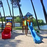 Plac zabaw nad jeziorem w Gołuchowie z kolorowymi zjeżdżalniami i tunelem. W tle widać wodę i drzewa.