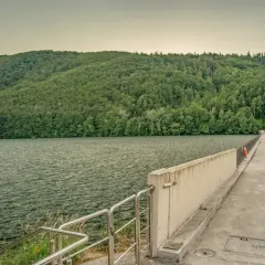 Zapora wodna z betonowym chodnikiem i balustradą, otoczona zielonymi wzgórzami i jeziorem.