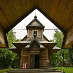 Drewniana cerkiew z krzyżami na tle zieleni. W oddali widać wiele krzyży, tworzących niezwykłą grabarkę.