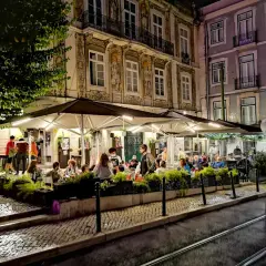 Wieczorny gwar w **Bairro Alto**. Ludzie siedzą przy stolikach pod parasolami, delektując się posiłkiem i atmosferą.