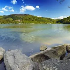 Kamienista plaża nad Jeziorem Międzybrodzkim, otoczona zielonymi wzgórzami i błękitnym niebem z białymi chmurami.