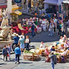 Tłoczny plac z fontanną i rzeźbą, kawiarnie z czerwonymi stolikami i ludźmi siedzącymi na zewnątrz, sklep z ubraniami w tle. Wszystko pod słońcem, tworząc wakacyjny, **coperto** nastrój.