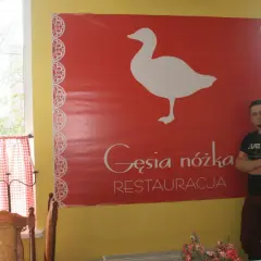 Mężczyzna stoi obok wielkiego czerwonego banera z białym gęsią i napisem "Gęsia nóżka RESTAURACJA".