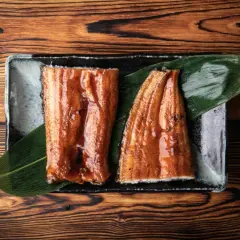 Dwa kawałki glazurowanego unagi na talerzu z liściem, na drewnianym tle.