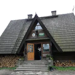 Drewniane schronisko Maciejowa z otwartymi drzwiami, stosy drewna po bokach i drabina na dachu.