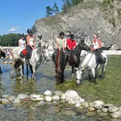 Konni jeźdźcy na koniach w rzece, w tle skaliste zbocze i plaża z ludźmi. To widok z Bukowiny Tatrzańskiej, jakby rynek atrakcji.