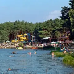 Tłum ludzi wypoczywa nad wodą, widać zjeżdżalnie wodne i drzewa. Krasnobród zalew tętni życiem w słoneczny dzień.