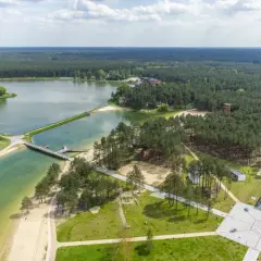 Malowniczy krajobraz z lotu ptaka: rozległy las otacza turkusowe wody zalewu janowskiego, z piaszczystymi plażami i nowoczesną architekturą.