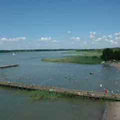 Wypływa z jeziora roś, a plażowicze kąpią się i opalają. W oddali widać żaglówki i łodzie.