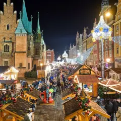 Tłoczny jarmark we Wrocławiu nocą, udekorowany świątecznymi światłami. Drewniane stragany oferują przysmaki, a tłumy spacerują wśród kolorowych kamienic.