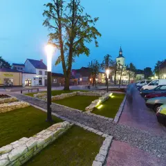 Rynek w Żarkach Letnisko o zmierzchu, z oświetlonymi latarniami, brukowanymi alejkami i zaparkowanymi samochodami.