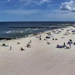 Słoneczny dzień na plaży w Dźwirzynie. Ludzie odpoczywają na piasku, kąpią się w morzu i spacerują po deptaku.