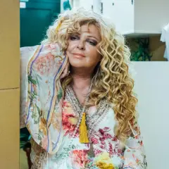 Kobieta z kręconymi blond włosami w kwiecistej sukience, opierająca się o ścianę.
