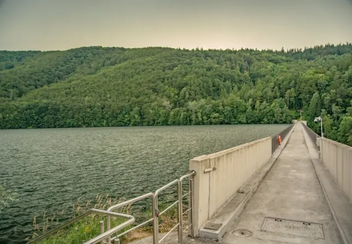 Zapora wodna z betonowym chodnikiem i balustradą, otoczona zielonymi wzgórzami i jeziorem.