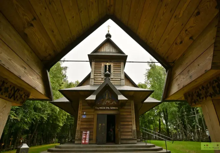 Drewniana cerkiew z krzyżami na tle zieleni. W oddali widać wiele krzyży, tworzących niezwykłą grabarkę.
