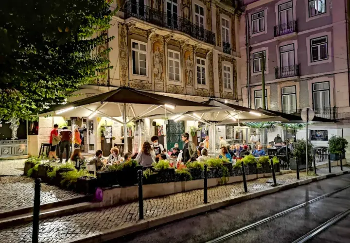 Wieczorny gwar w **Bairro Alto**. Ludzie siedzą przy stolikach pod parasolami, delektując się posiłkiem i atmosferą.