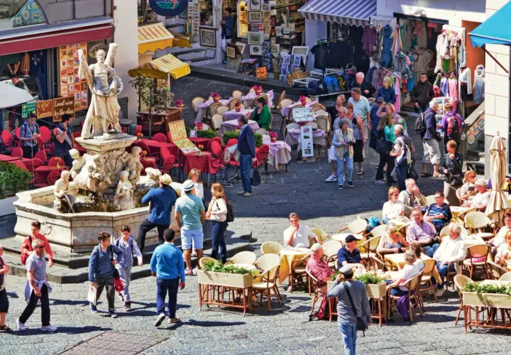 Tłoczny plac z fontanną i rzeźbą, kawiarnie z czerwonymi stolikami i ludźmi siedzącymi na zewnątrz, sklep z ubraniami w tle. Wszystko pod słońcem, tworząc wakacyjny, **coperto** nastrój.
