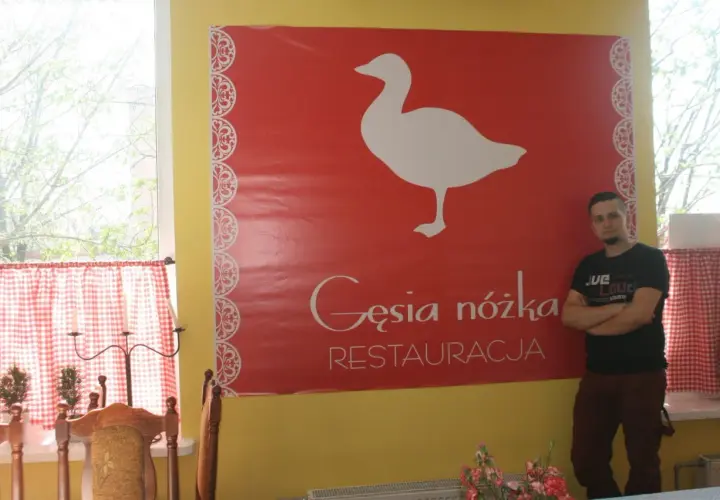 Mężczyzna stoi obok wielkiego czerwonego banera z białym gęsią i napisem "Gęsia nóżka RESTAURACJA".