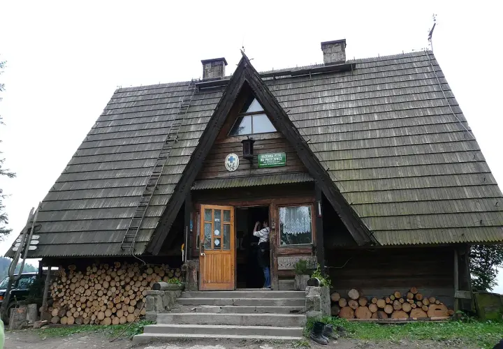 Drewniane schronisko Maciejowa z otwartymi drzwiami, stosy drewna po bokach i drabina na dachu.