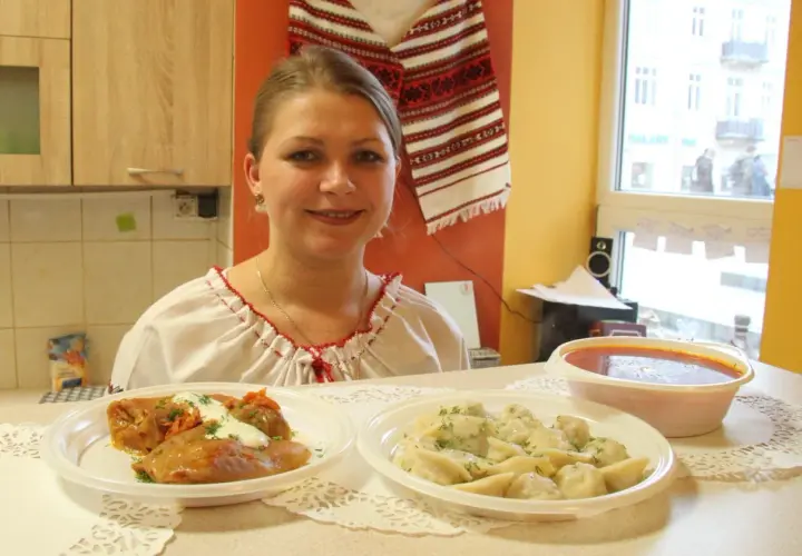 Uśmiechnięta kobieta w tradycyjnej koszuli prezentuje dania: gołąbki, pierogi i zupę. Pyszny, domowy obiad jak u Tarasa Bulby.