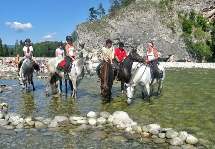 Konni jeźdźcy na koniach w rzece, w tle skaliste zbocze i plaża z ludźmi. To widok z Bukowiny Tatrzańskiej, jakby rynek atrakcji.