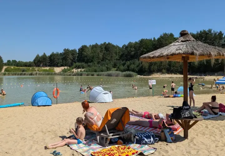 Słoneczny dzień nad zalewem Biszcza. Ludzie opalają się na plaży, kąpią w wodzie i odpoczywają pod parasolami.