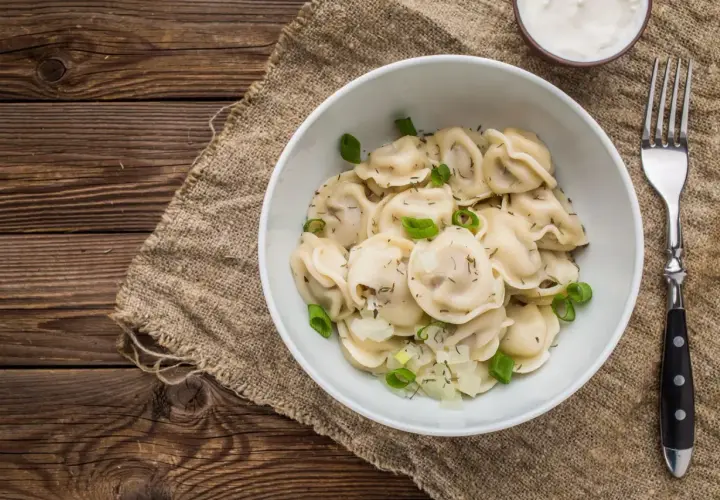 Pyszne pierogi z cebulką i koperkiem, idealne na obiad w stylu zapiecek warszawa. Obok miseczka śmietany i widelec.