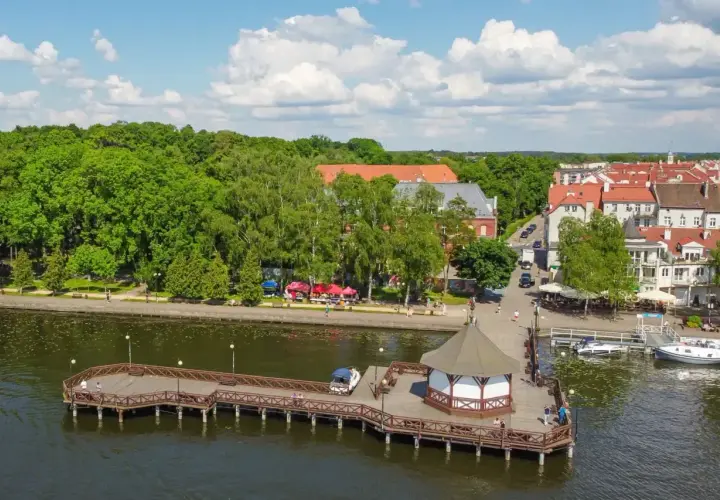 Malownicze miejscowości na Mazurach: drewniane molo z altaną, zielony park i urokliwe kamienice nad jeziorem.