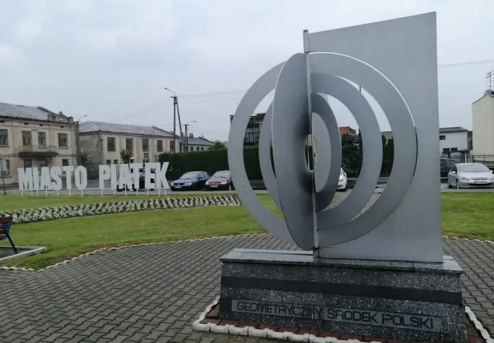 Geometryczny środek Polski w miejscowości Piątek. Monumentalna rzeźba i napis "Miasto Piątek" na tle budynków.