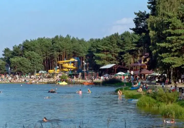 Tłum ludzi wypoczywa nad wodą, widać zjeżdżalnie wodne i drzewa. Krasnobród zalew tętni życiem w słoneczny dzień.