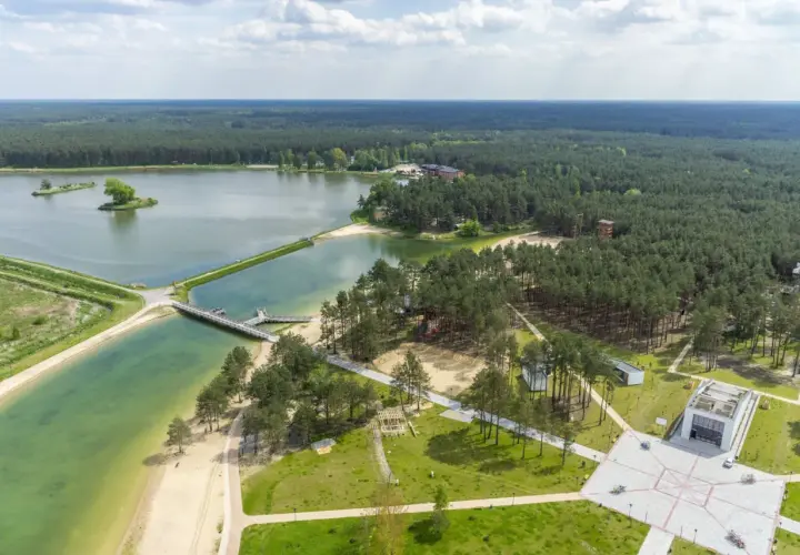 Malowniczy krajobraz z lotu ptaka: rozległy las otacza turkusowe wody zalewu janowskiego, z piaszczystymi plażami i nowoczesną architekturą.