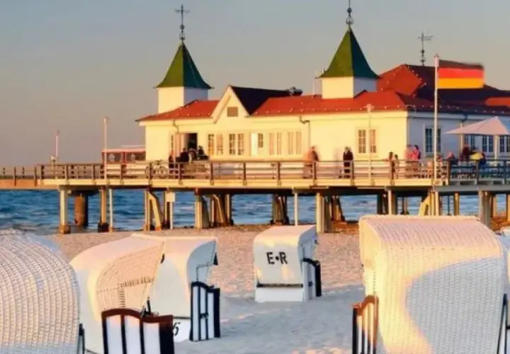 Zachód słońca nad molo w Ahlbecku. Białe kosze plażowe na piasku, a w tle budynek z restauracją i niemiecką flagą. To jedne z wielu atrakcji Ahlbecku.