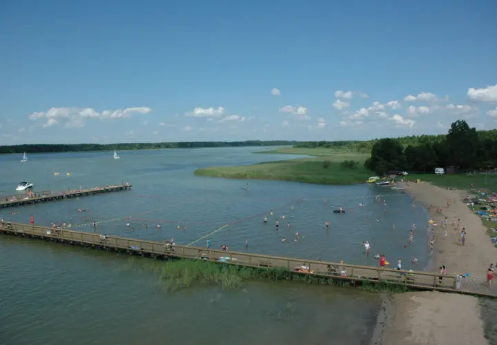 Wypływa z jeziora roś, a plażowicze kąpią się i opalają. W oddali widać żaglówki i łodzie.