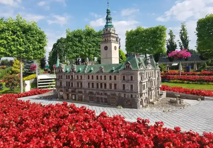 Miniatura ratusza z wieżą zegarową, otoczona czerwonymi kwiatami, w miniaturowej Kotlinie Kłodzkiej.