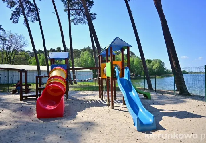 Plac zabaw nad jeziorem w Gołuchowie z kolorowymi zjeżdżalniami i tunelem. W tle widać wodę i drzewa.
