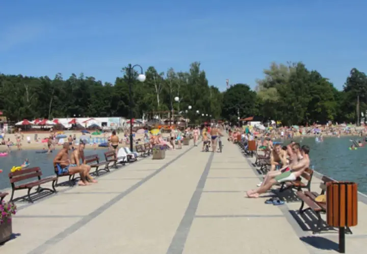 Ludzie odpoczywają na pomoście nad Skorzęcinem, jezioro tętni życiem, plaża pełna plażowiczów.