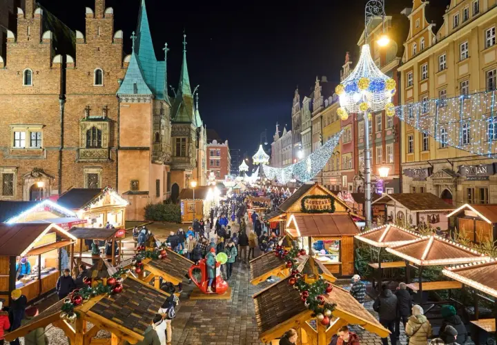 Tłoczny jarmark we Wrocławiu nocą, udekorowany świątecznymi światłami. Drewniane stragany oferują przysmaki, a tłumy spacerują wśród kolorowych kamienic.