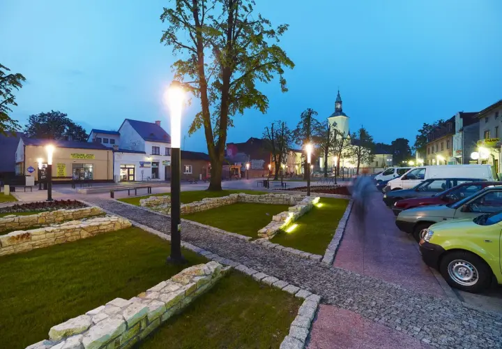 Rynek w Żarkach Letnisko o zmierzchu, z oświetlonymi latarniami, brukowanymi alejkami i zaparkowanymi samochodami.