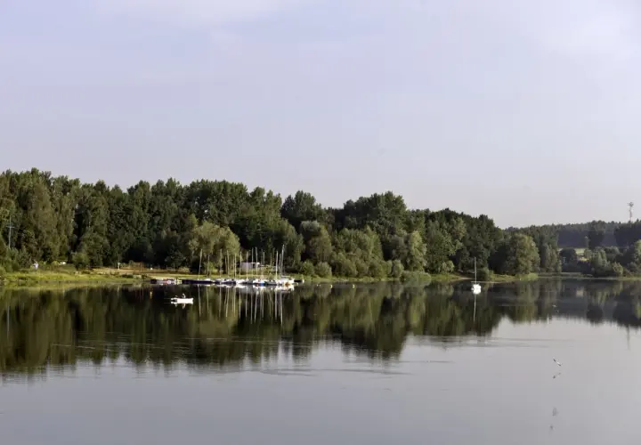 Zalew Przeczyce z łodziami przy brzegu i samotną białą żaglówką na spokojnej tafli wody.