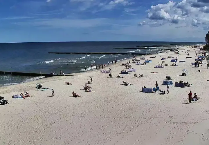 Słoneczny dzień na plaży w Dźwirzynie. Ludzie odpoczywają na piasku, kąpią się w morzu i spacerują po deptaku.