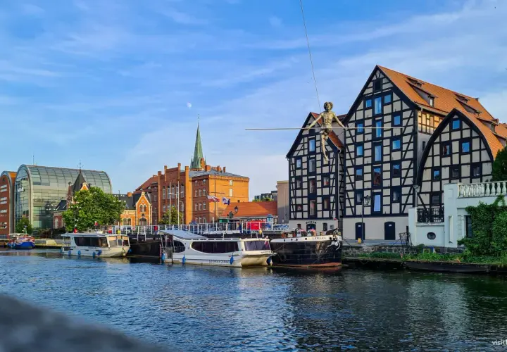 Bydgoszcz wydarzenia dzisiaj: na Wyspie Młyńskiej spaceruje po linie figura, cumują statki, a w tle widać zabytkowe kamienice i nowoczesną architekturę.