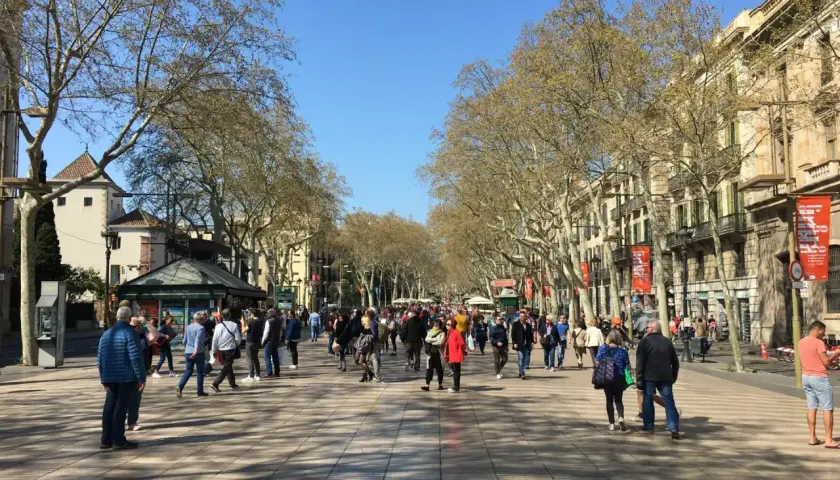Tłum ludzi spaceruje po La Rambla w Barcelonie, pod cienistymi drzewami i wzdłuż zabytkowych budynków.