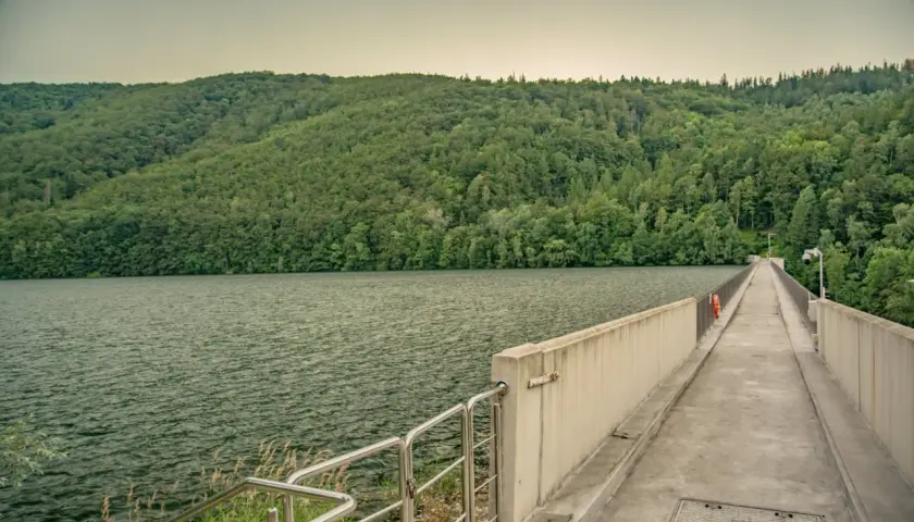 Zapora wodna z betonowym chodnikiem i balustradą, otoczona zielonymi wzgórzami i jeziorem.