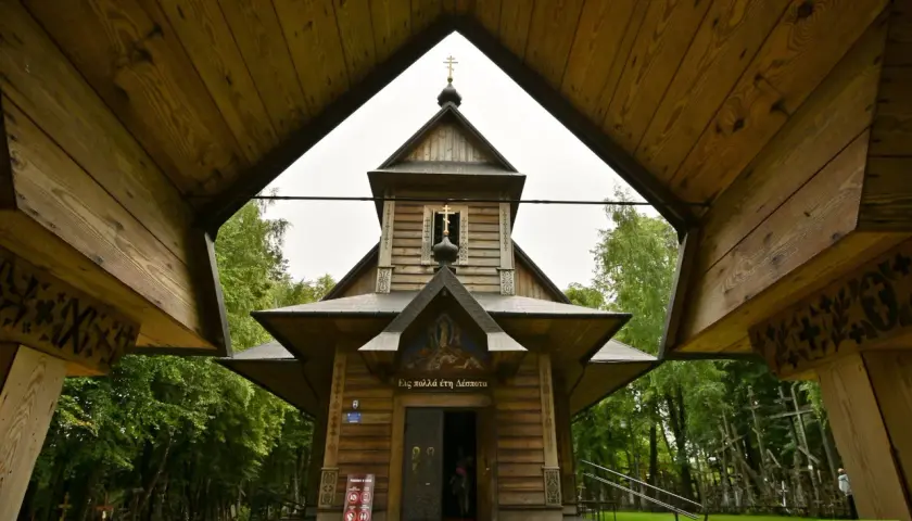 Drewniana cerkiew z krzyżami na tle zieleni. W oddali widać wiele krzyży, tworzących niezwykłą grabarkę.
