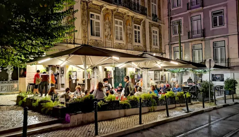 Wieczorny gwar w **Bairro Alto**. Ludzie siedzą przy stolikach pod parasolami, delektując się posiłkiem i atmosferą.