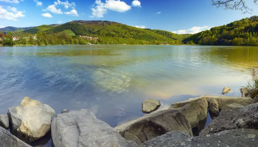 Kamienista plaża nad Jeziorem Międzybrodzkim, otoczona zielonymi wzgórzami i błękitnym niebem z białymi chmurami.