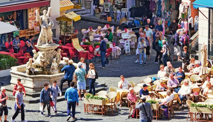 Tłoczny plac z fontanną i rzeźbą, kawiarnie z czerwonymi stolikami i ludźmi siedzącymi na zewnątrz, sklep z ubraniami w tle. Wszystko pod słońcem, tworząc wakacyjny, **coperto** nastrój.