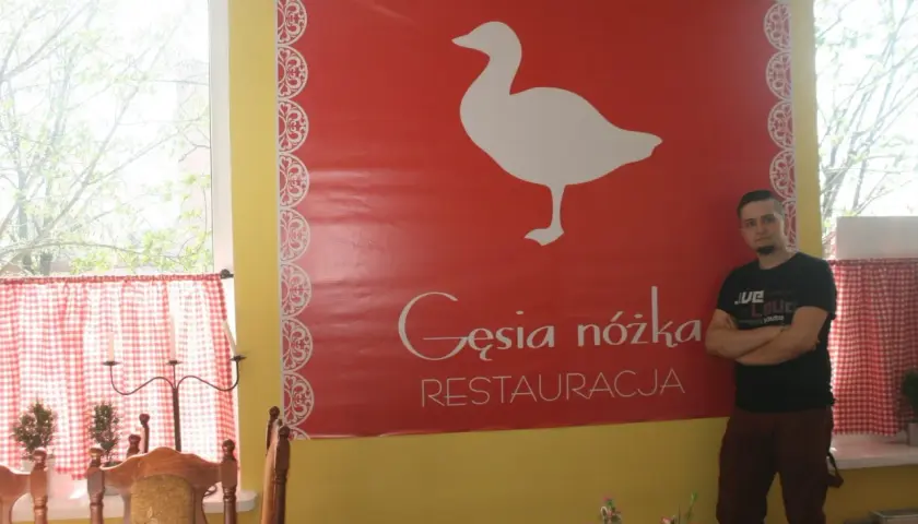 Mężczyzna stoi obok wielkiego czerwonego banera z białym gęsią i napisem "Gęsia nóżka RESTAURACJA".