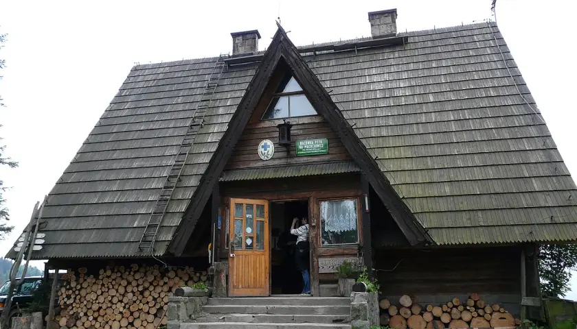 Drewniane schronisko Maciejowa z otwartymi drzwiami, stosy drewna po bokach i drabina na dachu.