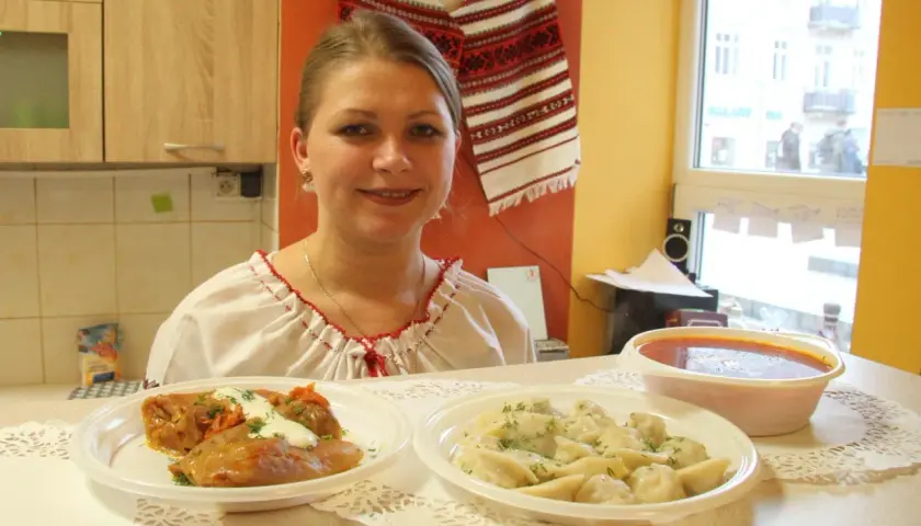 Uśmiechnięta kobieta w tradycyjnej koszuli prezentuje dania: gołąbki, pierogi i zupę. Pyszny, domowy obiad jak u Tarasa Bulby.
